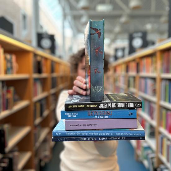 Ditte holder et udvalg af de nævnte bøger fra artiklen frem for sig. Bag hende ses biblioteksrummet med fyldte bogreoler. Baggrunden er sløret.