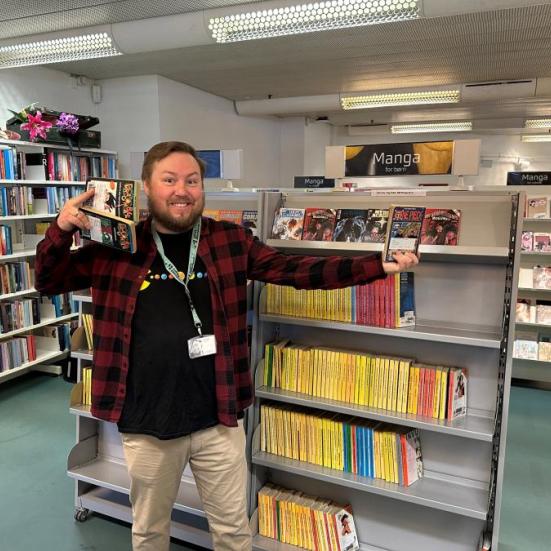 Kristian står smilende med tre mangaer i hænderne. Han står foran en bogreol fyldt med mangaer i Voksen Skønlitteratur på Hovedbiblioteket.