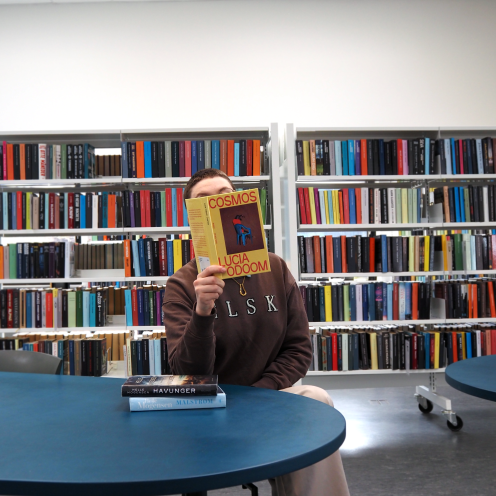 Anne Cecilie sidder ved et bord og læser i Lucia Odooms bog "Cosmos". På bordet foran hende ligger to af Helle Mogensens krimier med bogryggene vendt mod kameraet. Bag Anne Cecilie ses fyldte bogreoler og en solstråle.