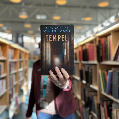 Hanne står mellem bogreoler og holder romanen "Tempel" frem for sig, så den dækker hendes ansigt. Der er fuld fokus på bogens forside og baggrunden er sløret. 