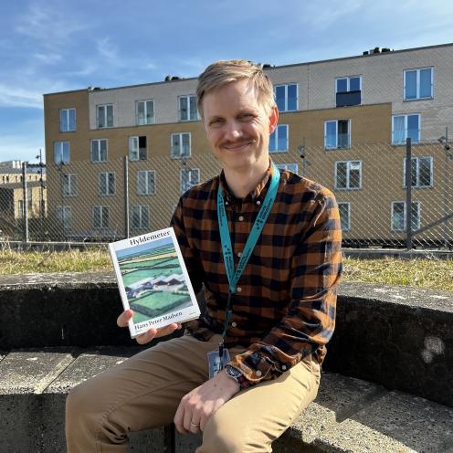 Mathias sidder i bibliotekshaven ved Hovedbiblioteket. Han holder bogen "Hyldemeter" i den ene hånd. Mathias smiler til kameraet. Himlen er blå og solen skinner.