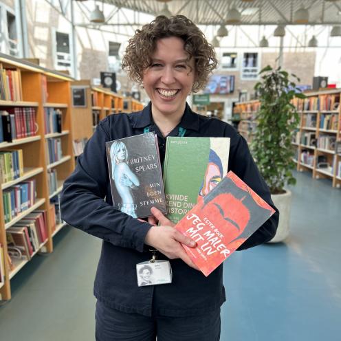 Ditte holder de tre omtalte bøger i denne artikel i favnen. Hun smiler stort til kameraet. Baggrunden, der viser resten af biblioteksrummet på Hovedbiblioteket, er let sløret.