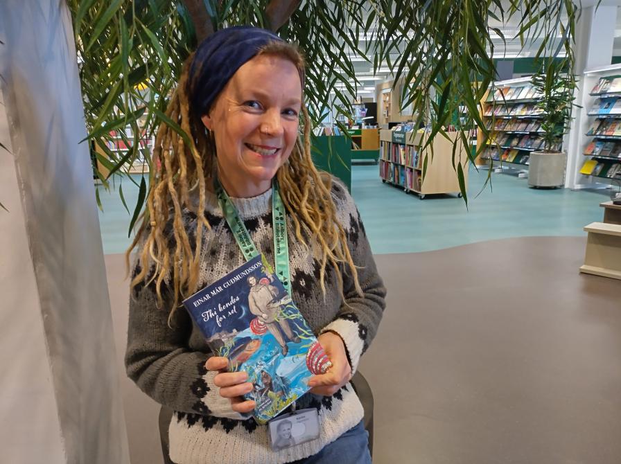 Karina står i afdelingen for skønlitteratur for voksne på Hovedbiblioteket. Hun holder "Thi kendes for ret" i hænderne og smiler til kameraet.