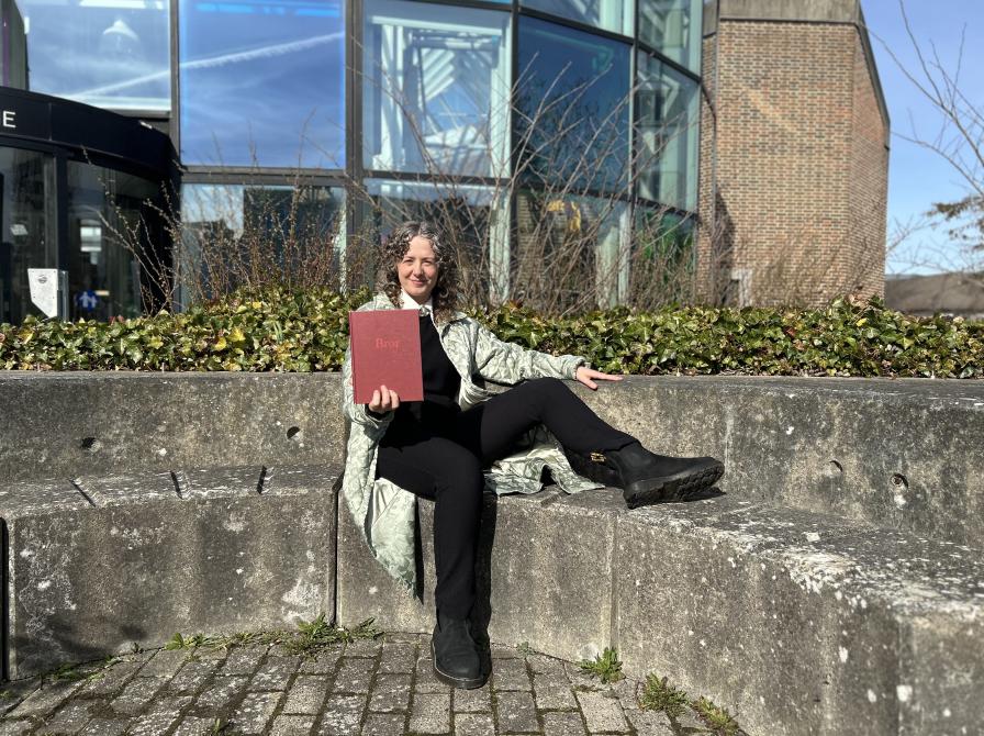 Majken sidder i bibliotekshaven ved Hovebiblioteket. Hun holder bogen "Bror" frem for sig og smiler. Bag Majken ses glashjørnets farverige vinduer. Himlen er blå.