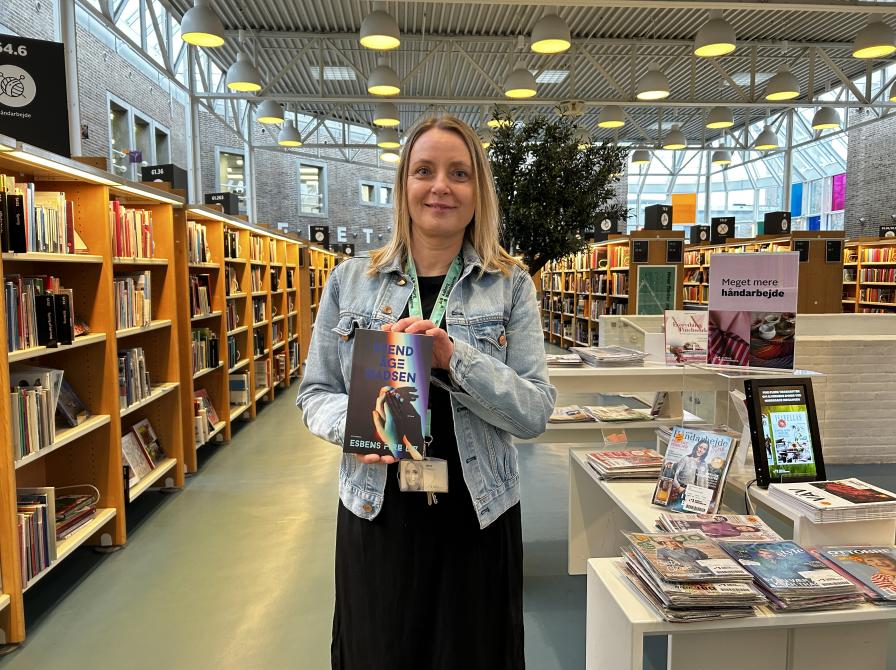 Nina står omgivet af bogreoler og udstillinger på Hovedbiblioteket. Hun holder "Esbens fire liv" i hænderne og smiler til kameraet.