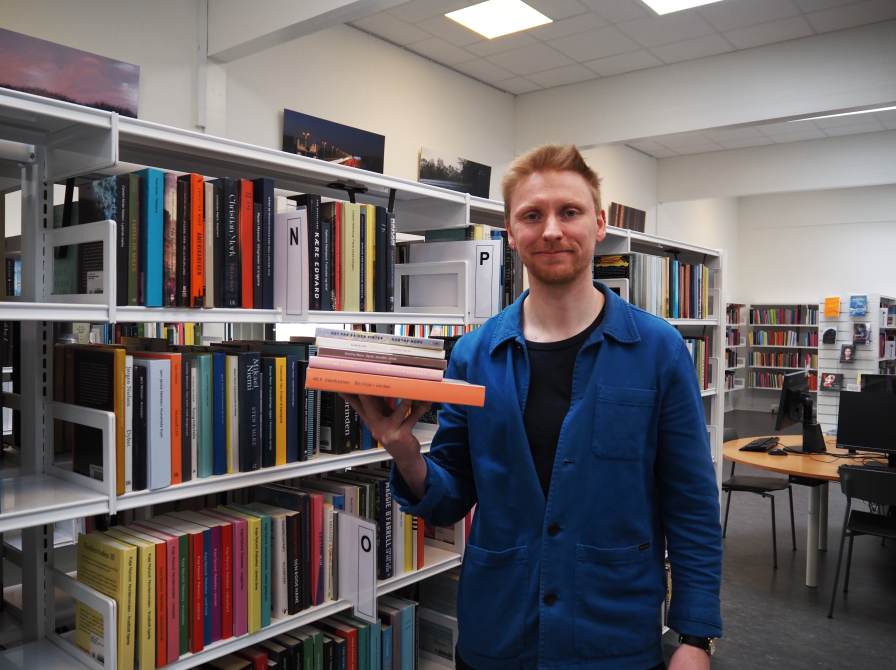 Patrick står med en stak af Dorthe Nors bøger. Bag ham ses biblioteksrummet med fyldte bogreoler. Han kigger ind i kameraet og smiler.