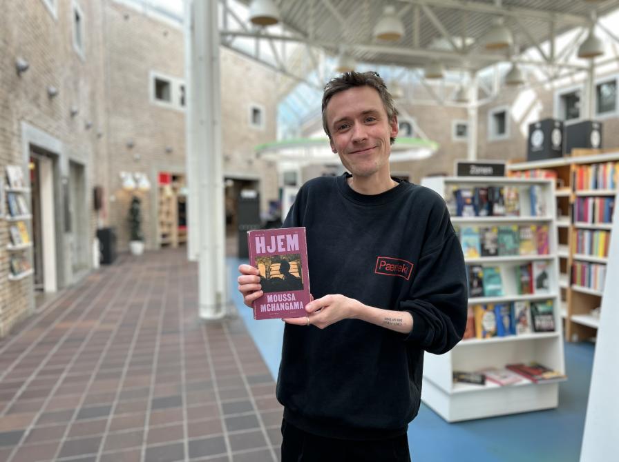 Søren Malthe står med den omtalte bog i denne artikel i hænderne. Han smiler til kameraet. Baggrunden er let sløret og viser biblioteksrummet.