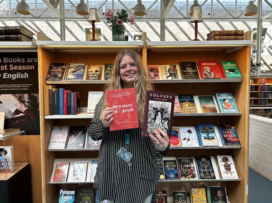 Sørine står foran en fyldt bogreol. Hun smiler og holder de to omtalte romaner fra denne artikel i hænderne.