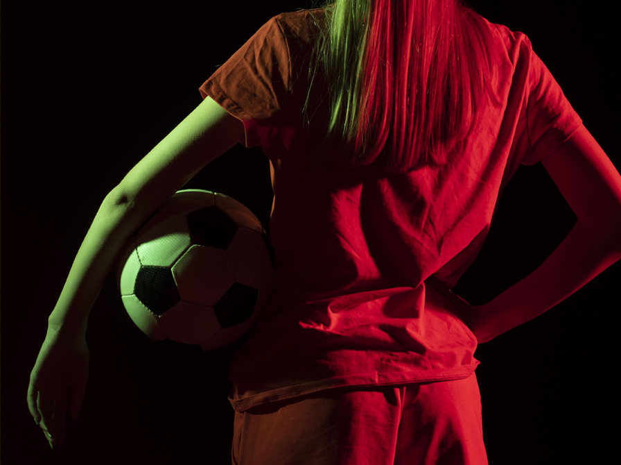 En kvindelig fodboldspiller med en bold under armen står med ryggen til.